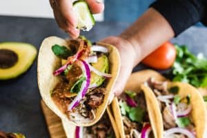 hands preparing mexican tacos with pork carnitas avocado onion cilantro and red sauce
