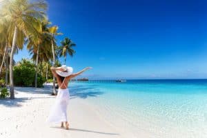woman with white hat walks down a tropical paradise beach with palm trees and turquoise sea
