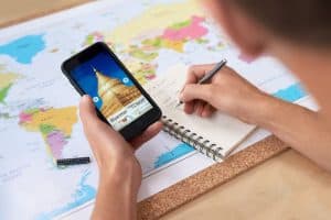 person writing travel plan and holding mobile phone at table with map