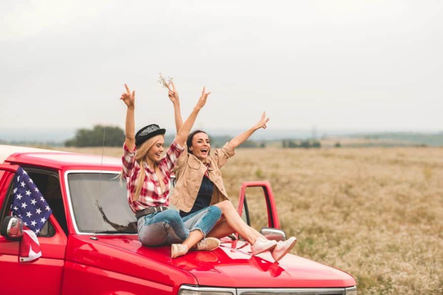 Beautiful young girlfriends sitting on car hood with raised hands and showing peace signs