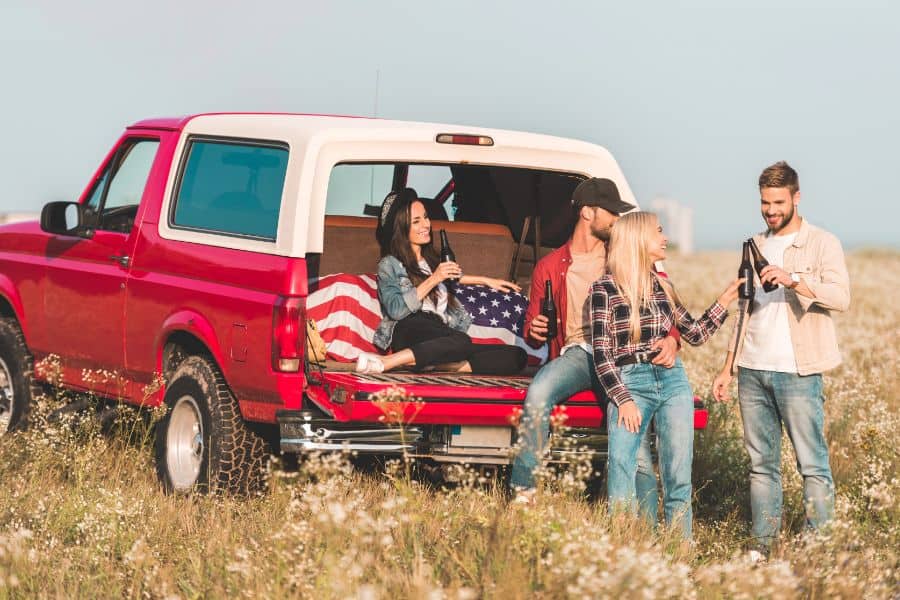 Group of young people clinking bottles of beer while sitting in car trunk