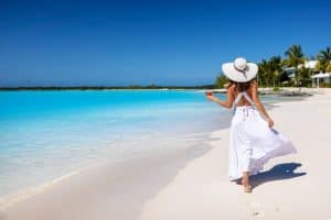 woman in a white summer dress and holding a drink walks along a tropical beach